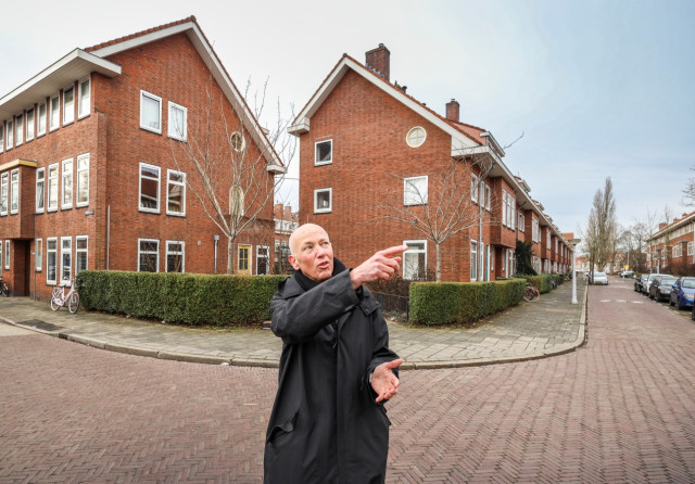 Witte man met kaal hoofd en lange jas wijst naar iets op straat.