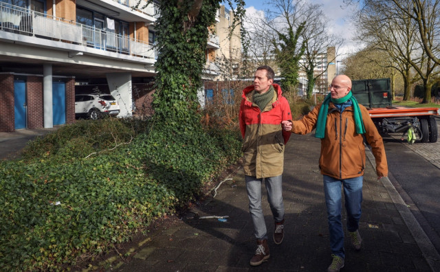 Twee witte mannen lopen door een wijk met links een flat en rechts een geparkeerd stuk vrachtwagen. Het is koud en ze dragen sjaals