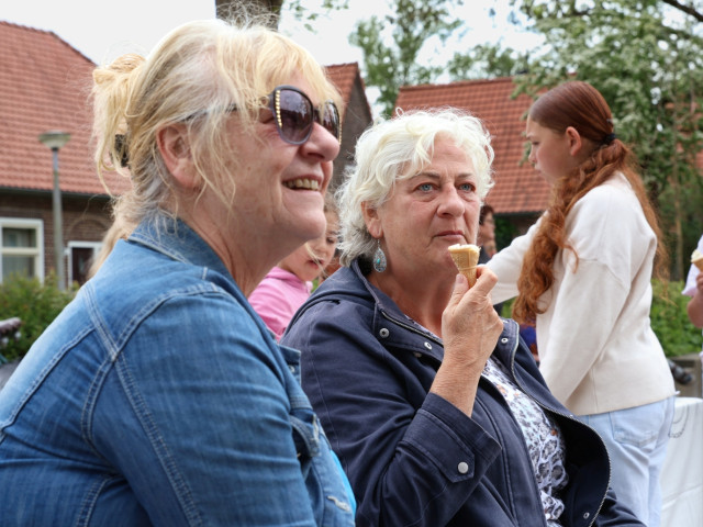 Twee witte vrouwen van een zekere leeftijd zitten op een pleintje, eentje eet een ijsje. Op de achtergrond zijn kinderen