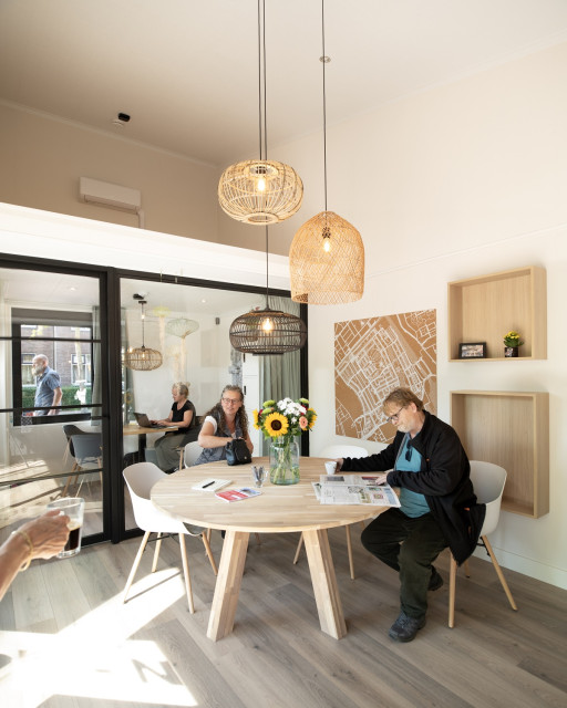 Witte mensen in een buurtgebouwtje. Eentje leest de krant, eentje pakt haar tas, de ander werkt op een laptop. Er staan zonnebloemen op tafel.