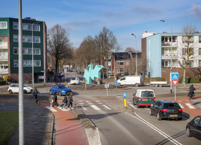 Een rotonde met veel autoverkeer, voetgangers en fietsers met daar omheen portiekwoningen