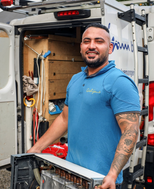 Man met tattoos en baardje draagt een blauwe polo van Gaslijn en heeft een ketel of warmtepompboiler in zijn handen. Op de achtergrond een witte werkbus met gereedschap.
