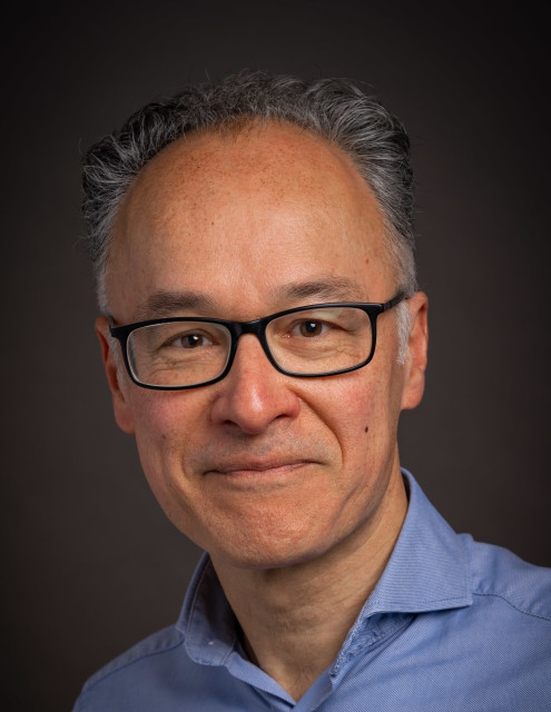 Witte man met grijs haar en bril. Hij lacht met zijn mond dicht en draagt een overhemd.