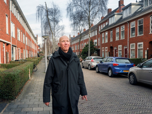 Witte man met kaal hoofd in lange donkere jas loopt over straat. Aan een kant zijn heggen aan de andere kant geparkeerde auto's te zien.