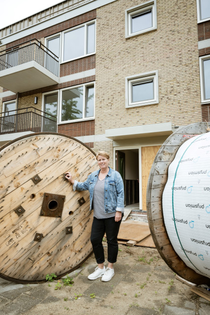 Witte vrouw met spijkerjasje en kort pittig kapsel bij een grote rol bouwmateriaal voor een nieuwbouwflat.