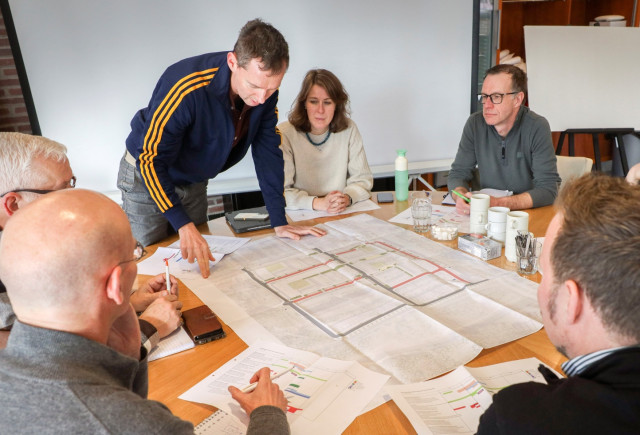 Een vergadertafel met kaarten en een hoop witte mensen er omheen