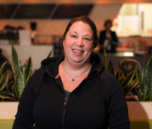 Witte vrouw met donkere capuchontrui vanuit een buurtcentrum met op de achtergrond een koffiebar en planten.