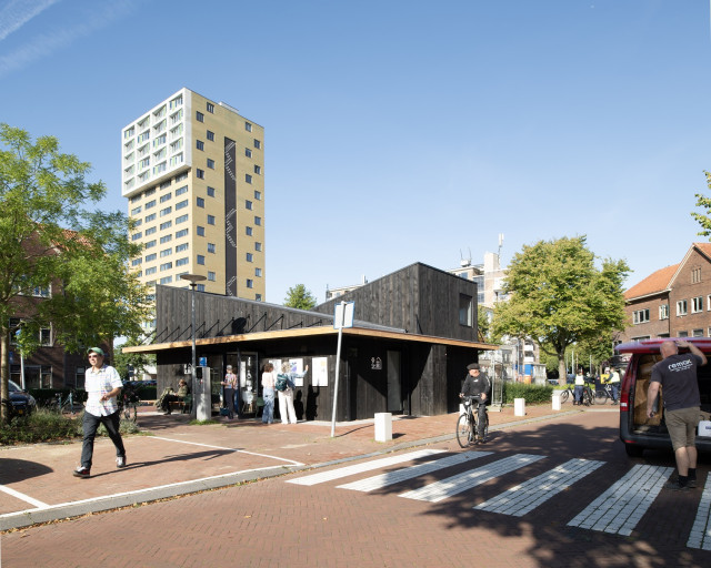 Een houten wijkgebouwtje op een plein. Op de voorgrond een zebrapad en een fietser.
