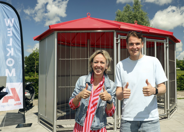 Een witte vrouw en een witte man lachen allebei in de camera en steken hun duimen omhoog. Op de achtergrond een nieuwe hangplek voor jongeren en een grote beachflag van Alfa-college met het woord WELKOM erop.
