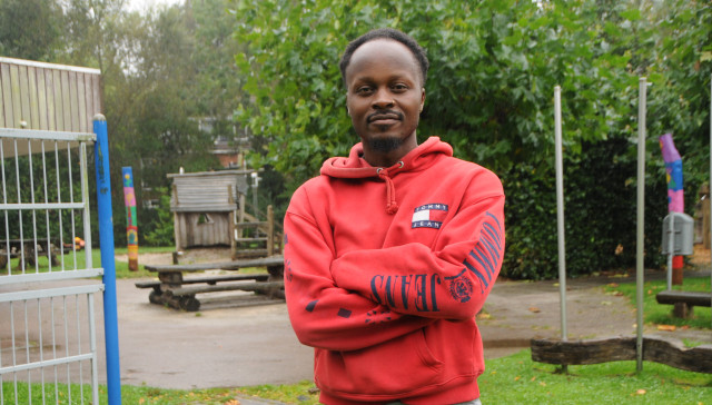 Donkere man met een rode hoodie en de armen over elkaar bij een speelveldje