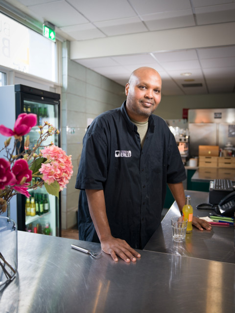 Zwarte man met kaal hoofd glimlacht. Hij staat achter een counter met een kassa en een flesje drinken.