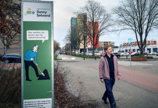 Witte man met snor loopt over een stoep met links een informatiebord en op de achtergrond een winkelcentrum.