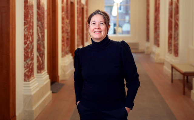 Witte vrouw met zwarte coltrui aan en handen in de zakken staat in de gang van het stadhuis in Groningen. Er is marmer en loper te zien