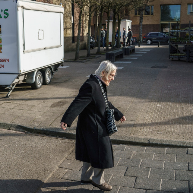 Oudere vrouw met lange jas en grijs haar loopt over straat, op de achtergrond een snackwagen
