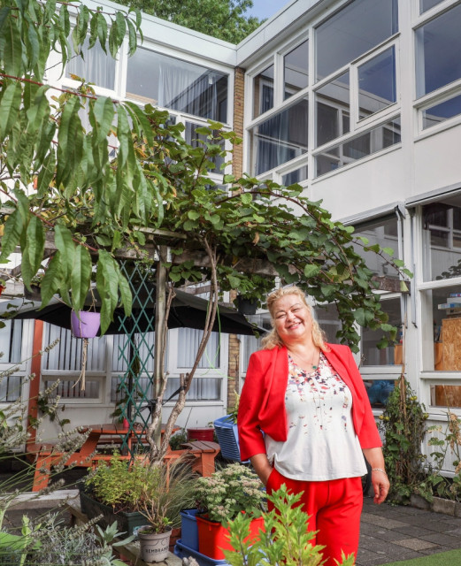 Vrouw met blond geverfd haar in een wit shirt en rood pak staat lachend in een tuin.