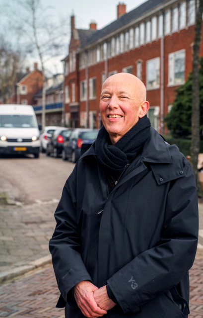 Witte man met kaal hoofd lacht in de camera. Hij heeft een lange jas en een sjaal om.