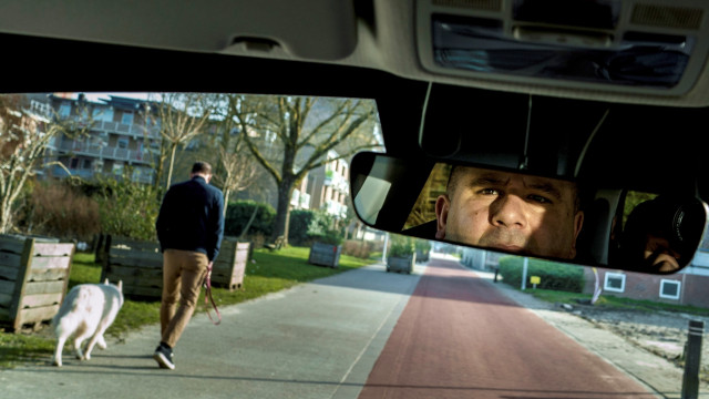 Uitzicht vanuit een auto. Een man kijkt in de achteruitkijkspiegel. Op straat is een fietspad en iemand die een hond uitlaat.