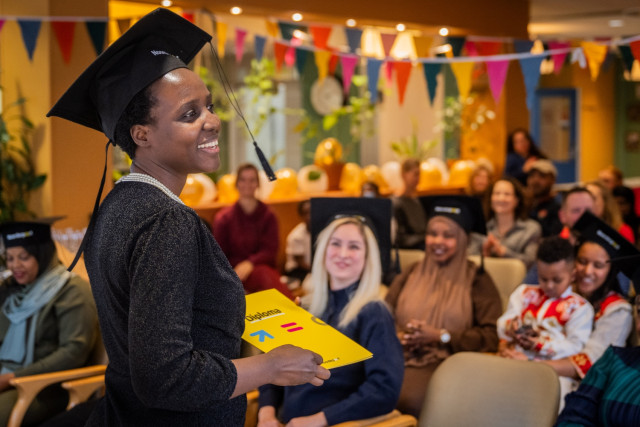 Zwarte vrouw met diploma in haar hand en afstudeerhoed op lachend voor een zaal met toeschouwers en vlaggetjes