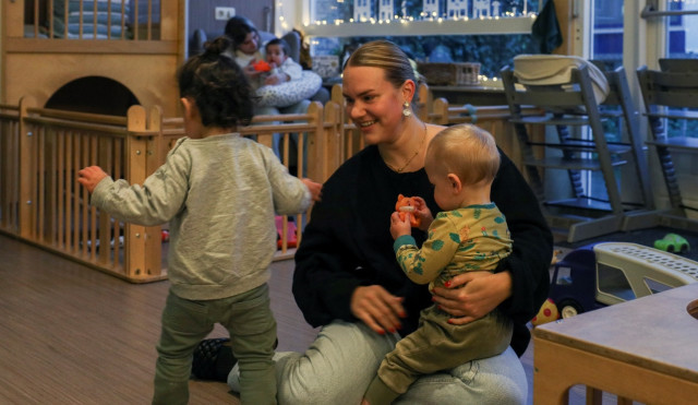 Witte vrouw met blond haar en zwarte trui zit op de grond. Op haar schoot een baby en daarnaast een iets groter kindje. Ze zitten in een kinderopvang met allemaal speelgoed en kinderstoelen.