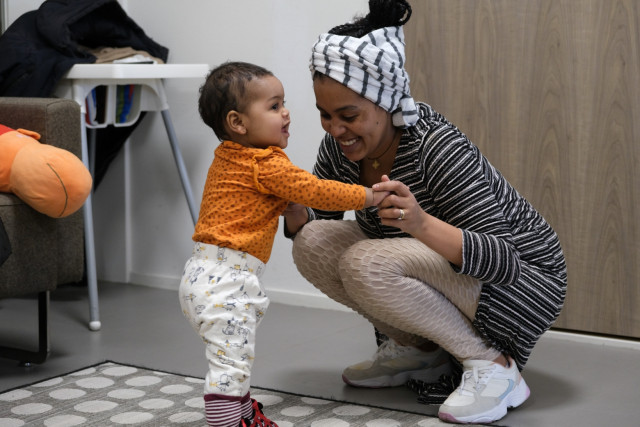 Lichtgetinte vrouw zit op haar hurken en houdt een klein kindje vast die probeert te lopen
