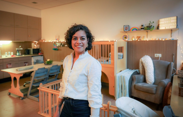 Vrouw met donker krullend haar en een wit shirt staat lachend voor de camera. Ze staat in een ruimte van de kinderopvang. Met een keukentje, kinderstoelen en bedjes.