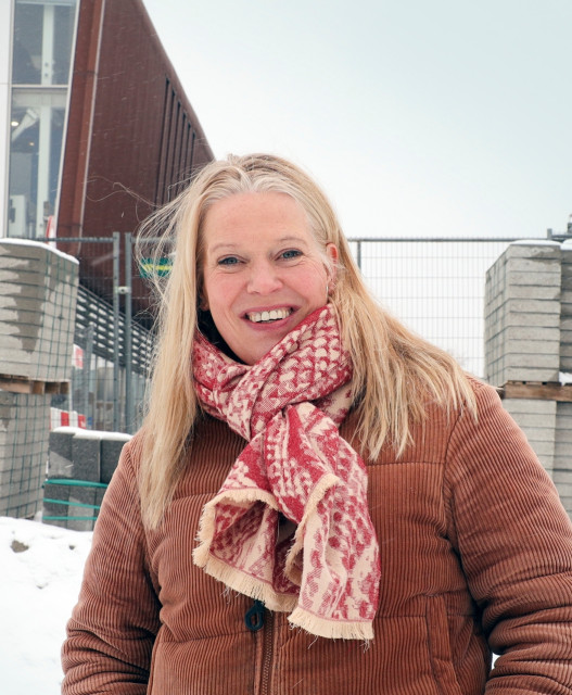Witte vrouw met blond haar en gekleurde sjaal staat in een winters landschap waar wordt gewerkt. Er zijn bouwhekken en bakstenen.