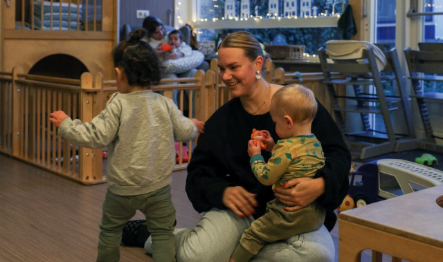 Witte vrouw met blond haar en zwarte trui zit op de grond. Op haar schoot een baby en daarnaast een iets groter kindje. Ze zitten in een kinderopvang met allemaal speelgoed en kinderstoelen.
