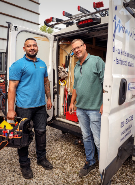 Een lichtgetinte man en een witte man staan samen voor een werkbus met gereedschap. De ene man draagt een gereedschapskist.
