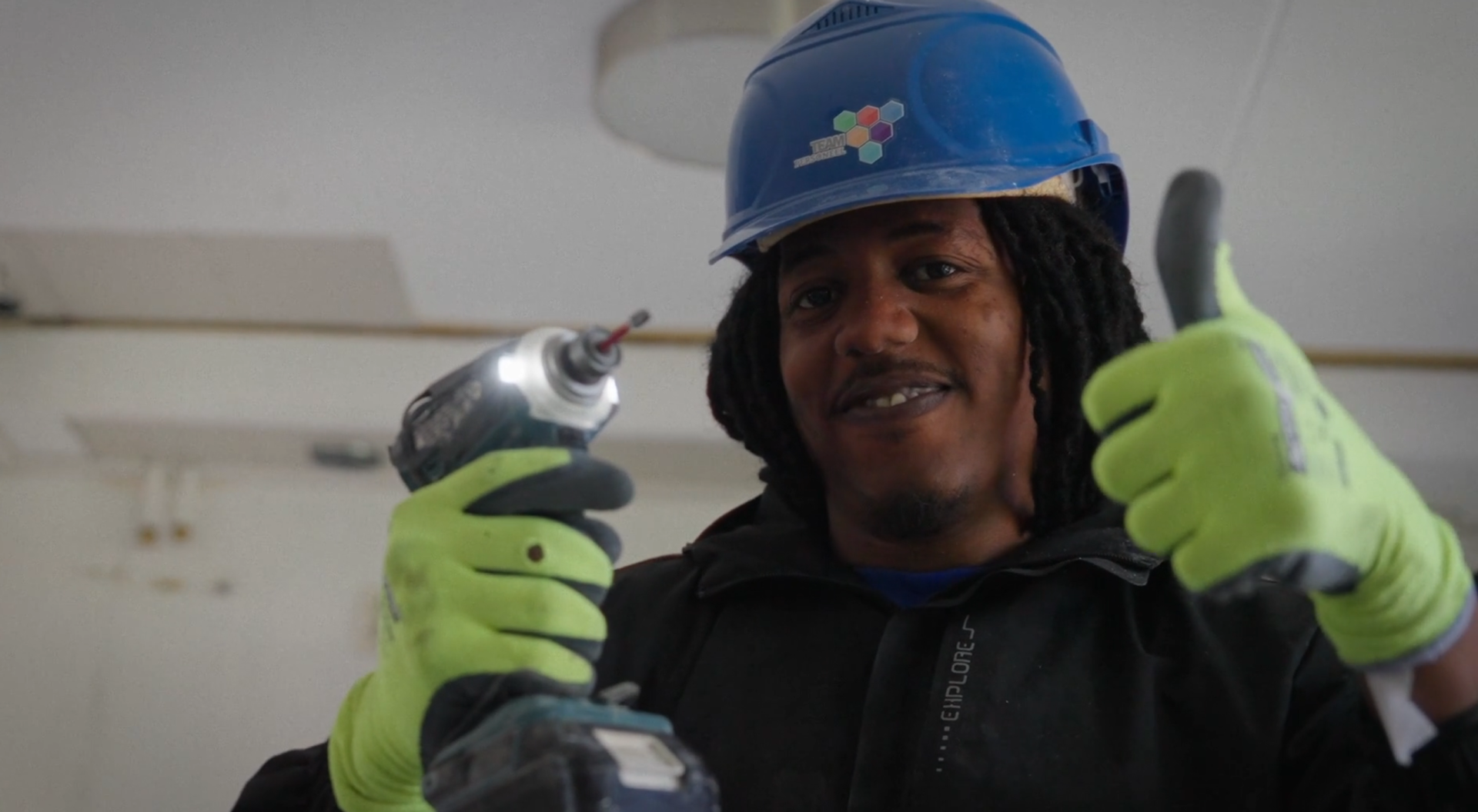 De AanDeBak-garantie film over Jonathan. Een still uit de video over dit project. Daarop is een donkere man te zien met lang haar. Hij draagt een bouwvakkershelm en werkhandschoenen. Hij heeft een boormachine in zijn ene hand en steekt zijn duim op met zijn andere hand.