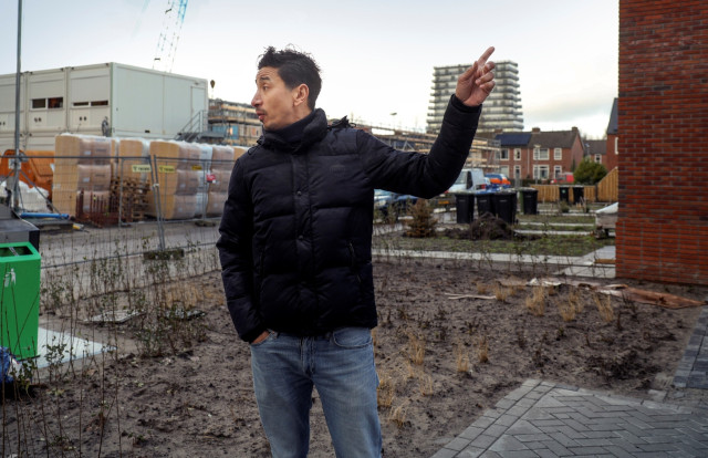 Man met donkere jas en spijkerbroek loopt door een wijk waar gebouwd wordt en wijst wat aan terwijl hij naar mensen uit beeld kijkt