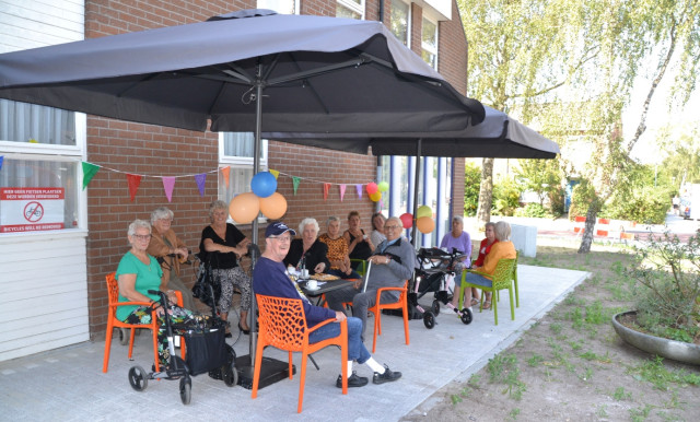 Een groep oudere, witte mensen zit buiten op tuinstoelen onder parasols. Er hangen gekleurde vlaggetjes aan de muur. Er staan een paar rollators.