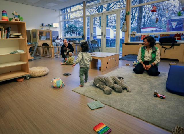 Ruimte in een kinderopvang. Met een paar kleine baby's en jonge kinderen. Twee begeleiders zitten er op de grond bij. Er ligt speelgoed op de groen en er zijn klimdingen.