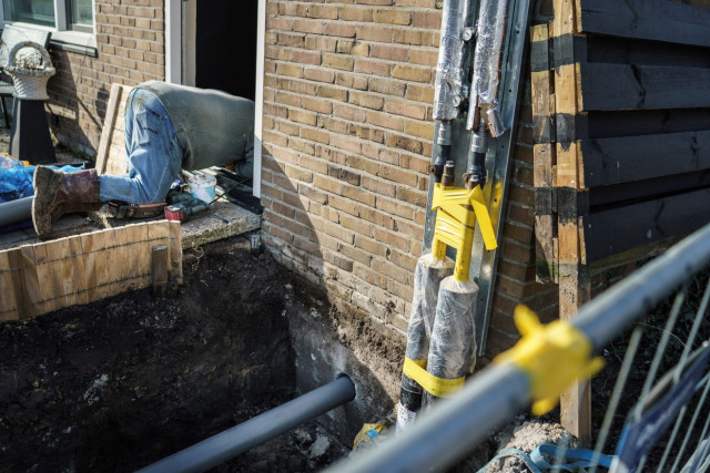 Bouwmateriaal en een tuin die open ligt. Uit een deuropening steekt een voorovergebukt persoon.
